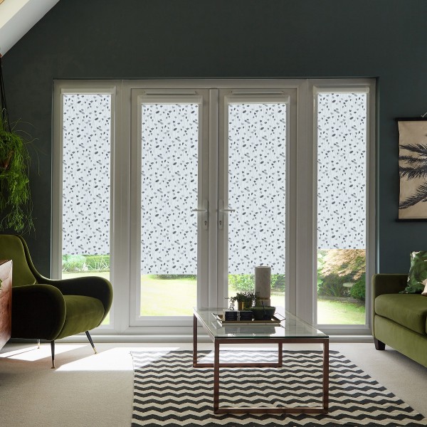 Glass double doors with patterned blinds blocking and diffusing daylight, in a modern living room with green armchairs, glass coffee table, chevron rug, and a view of the garden.