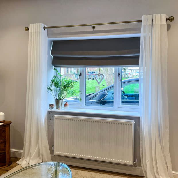 White sheer grommet curtains hang open on a brass rod, framing a folded grey Roman blind partially lowered; daylight filters into a living room with windowsill plant and radiator below.