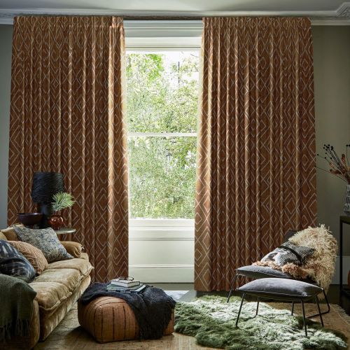 Patterned rust-orange, floor-length pleated curtains framing a sash window, pulled slightly apart to reveal bright greenery; they filter soft daylight into a cozy living room with plush sofa, pouf, chair.
