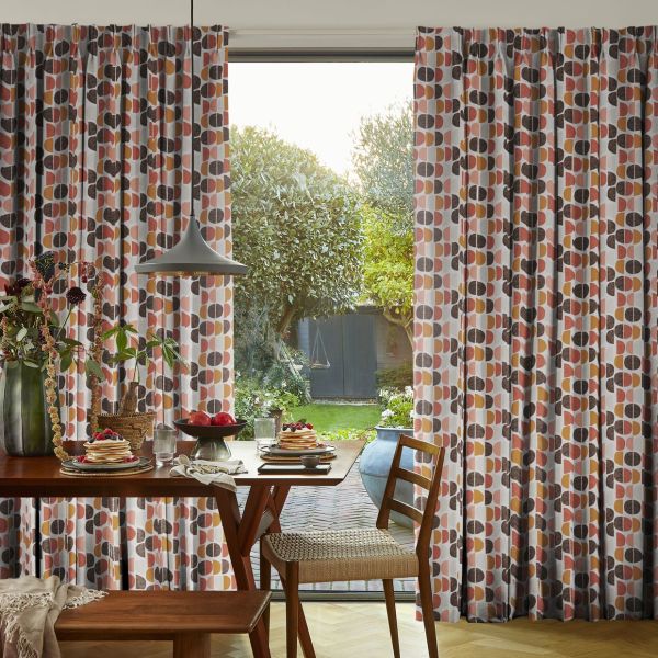 Full-length pleated patterned curtains (semi-circle design, orange, pink, brown) drawn to sides, framing a central opening that lets soft daylight through; dining table and pendant light face a green backyard.