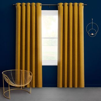 Mustard-gold grommet-top curtains, floor-length on a metal rod, are partially open, filtering bright daylight through a window against a deep navy wall with a gold wire chair and circular sconce.