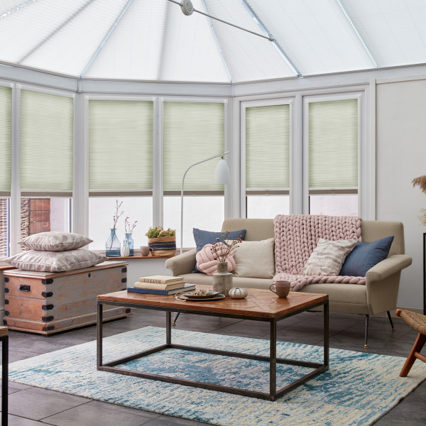 Light green pleated cellular blinds across tall angled conservatory windows, lowered to mid-height and softly filtering daylight; a bright seating area with sofa, wooden coffee table, rug, and plants.