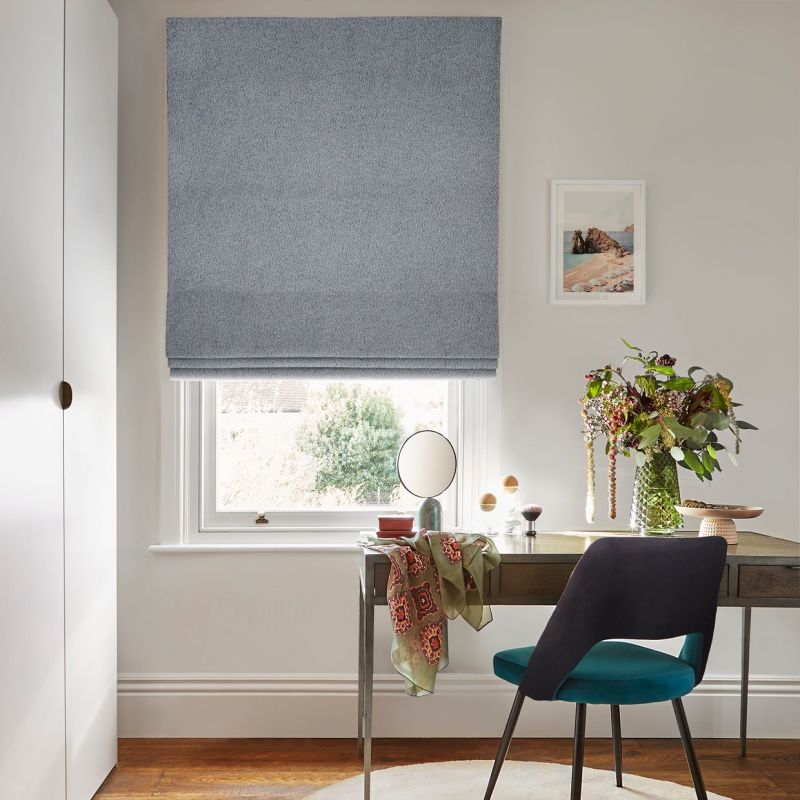 A textured light-blue Roman blind, lowered to mid-window with soft folds, gently filtering daylight; in a bright home office, desk, teal chair, vase of flowers and framed coastal print nearby.
