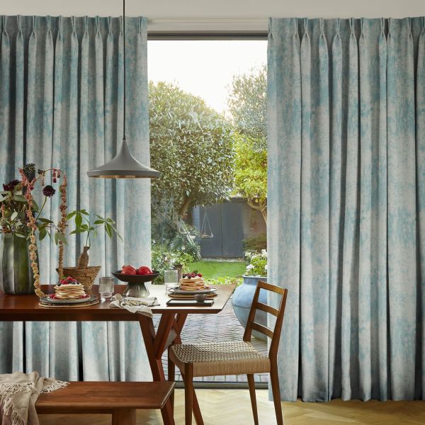 Full-length pleated blue-grey curtains, hanging on either side with soft folds — parted at the center, filtering morning light — framing a dining table set for breakfast and a sunlit garden.