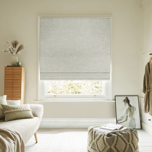 Light-gray textured Roman blind lowered with two soft folds, filtering diffuse daylight; in a neutral, cozy living room with sofa, wooden chest, knitted pouf and a leaning framed painting.