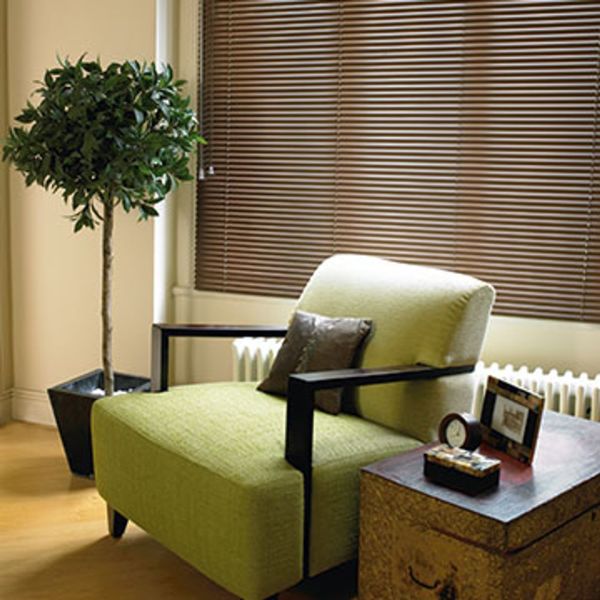 Brown venetian blinds cover the window, closed and softly filtering daylight; in a cozy living-room corner with a green armchair, trunk side table, potted topiary and hardwood floor.