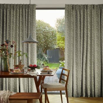 Full-length patterned curtains, beige with green fern print, parted to reveal sliding door; filtering soft daylight into a dining nook with wooden table set for breakfast and garden views.
