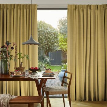 Floor-length pleated beige curtains, slightly parted at a sliding door, filtering daylight; wooden dining table set with pancakes and fruit beneath a pendant lamp, opening onto a lush backyard.