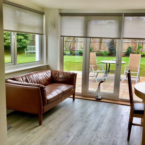 Light-grey roller blinds, lowered halfway across two windows and a sliding glass door, gently filtering daylight; overlooking patio with table and chairs, leather sofa in bright living-dining room.