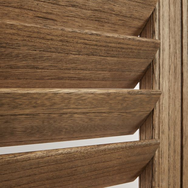 Louvered wooden shutters with wide horizontal slats, angled slightly open, showing pronounced wood grain and beveled edges, close-up interior view with warm tones and light through narrow gaps.