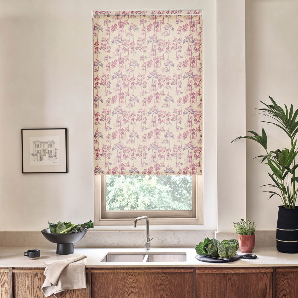 Floral-patterned roller blind hangs down over a kitchen window, filtering daylight; stainless-steel faucet and sink below, countertop with bowl of greens, potted plants, and a framed sketch nearby.