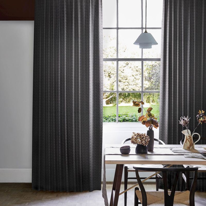 Dark grey, floor-length pleated curtains, hung at window sides with subtle vertical texture, partially drawn and filtering light, framing a sunlit dining room with table, chairs, vases and garden view.