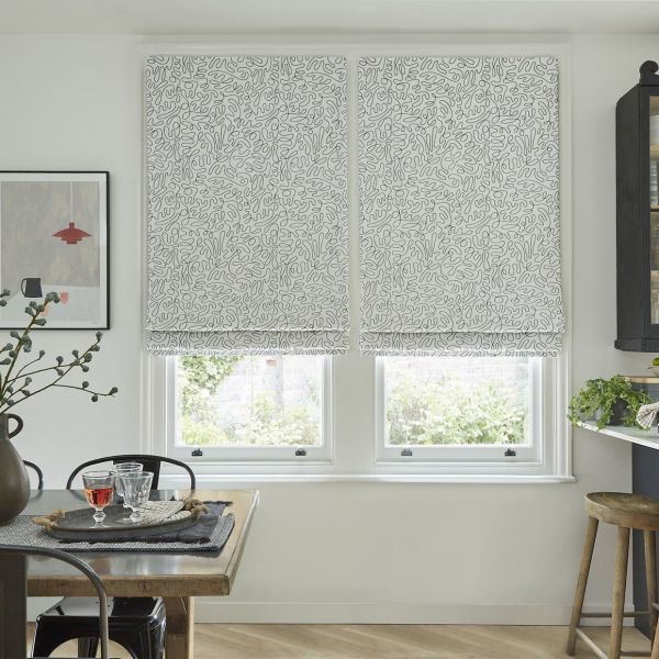 Patterned Roman blinds, lowered across two adjacent windows with an abstract squiggle print, filtering soft daylight; a casual dining/kitchen nook with wooden table, glasses, plants, framed art and stool.