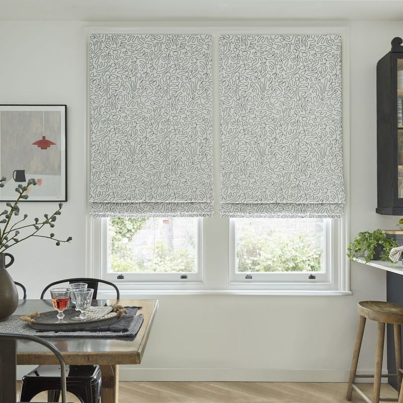 Patterned Roman blinds, lowered across two adjacent windows with an abstract squiggle print, filtering soft daylight; a casual dining/kitchen nook with wooden table, glasses, plants, framed art and stool.