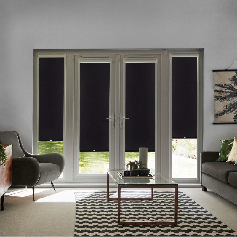 Black roller blinds on French doors and sidelights, mostly lowered blocking light; living room with grey armchair, sofa, glass coffee table, chevron rug, small garden glimpse.