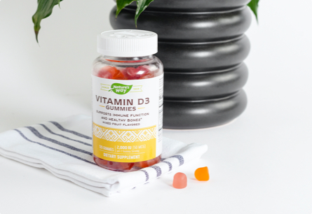 A bottle of Vitamin D3 Gummies sitting on top of a kitchen towel next to two orange gummies with a black potted plant in the background.