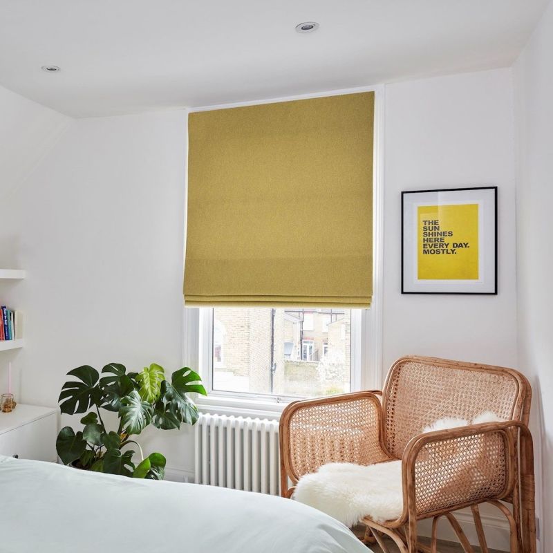Mustard-yellow Roman blind (lowered, slightly folded at bottom) filtering daylight. Bedroom corner with white walls, rattan armchair topped with sheepskin, monstera plant and a white radiator.
THE SUN SHINES HERE EVERY DAY. MOSTLY.
