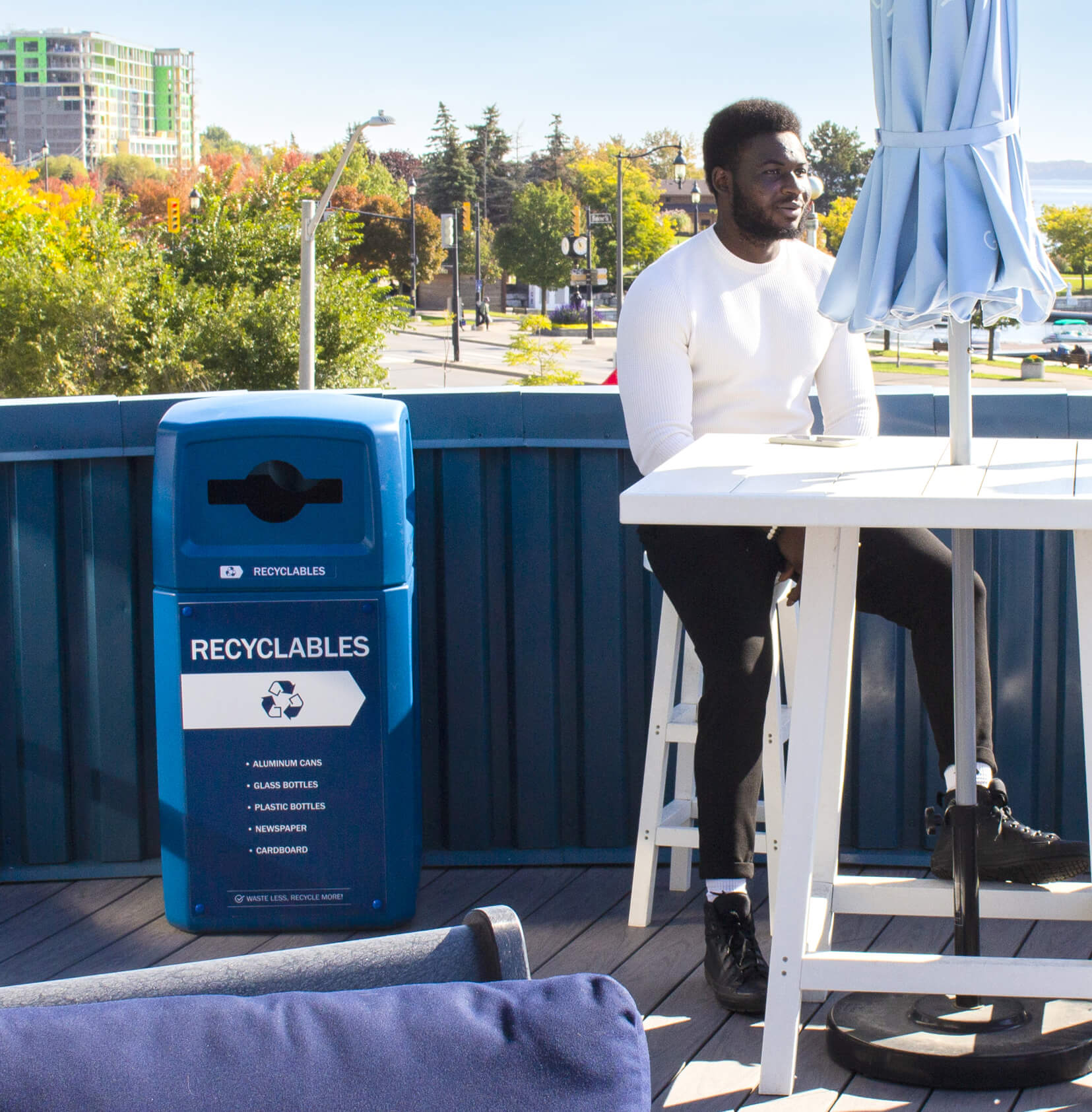 blue outdoor recycled plastic recyclables container with canopy lid and body sign outside on a rooftop patio with a man sitting at the table