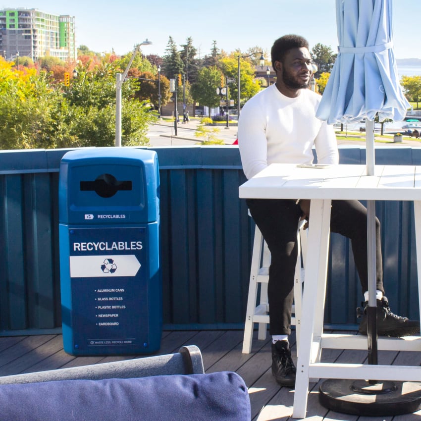 blue outdoor recycled plastic recyclables container with canopy lid and body sign outside on a rooftop patio with a man sitting at the table
