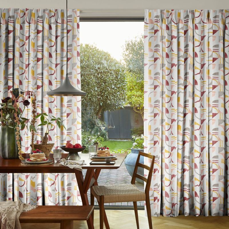 Pleated floor-length patterned curtains (pink, mustard, grey geometric print) pulled to sides, framing a glass door; filtering soft morning light over a breakfast table, pendant lamp, and garden view.