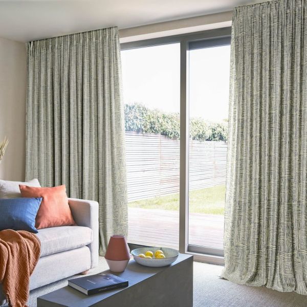 Patterned, floor-to-ceiling pleated curtains, drawn partly open at a sliding glass door, filtering bright daylight into a cozy living room with sofa, cushions, coffee table and a fenced backyard view.