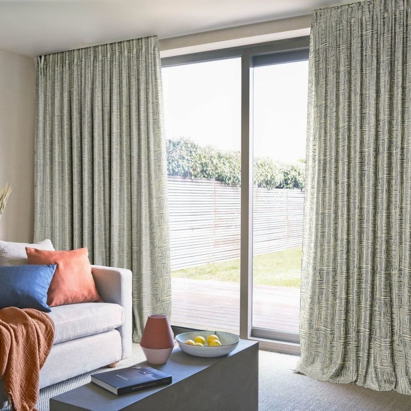 Patterned, floor-to-ceiling pleated curtains, drawn partly open at a sliding glass door, filtering bright daylight into a cozy living room with sofa, cushions, coffee table and a fenced backyard view.