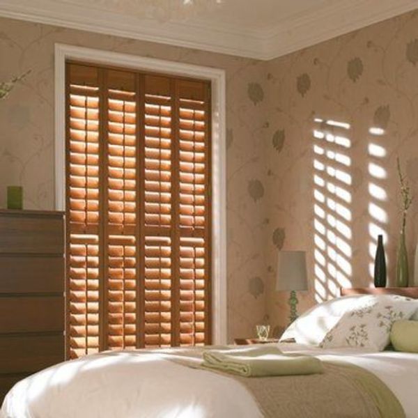 Wooden plantation shutters covering a tall window—four full-height panels with adjustable slats—partially closed, casting striped sunlight across a softly lit bedroom with bed, nightstand, patterned wallpaper and dresser.