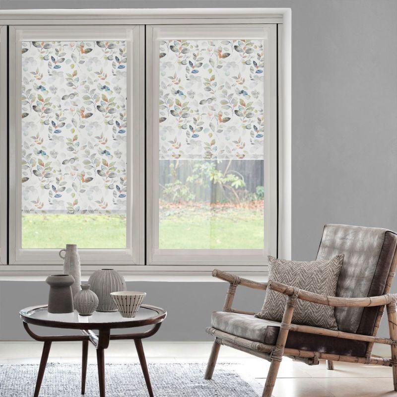 Object: floral roller blinds with muted watercolor leaves, mounted on twin windows and lowered halfway. Action: filtering daylight. Context: gray living room with rattan armchair, round table, and lawn view.