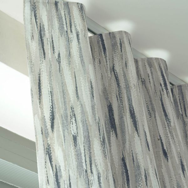Patterned wave-fold curtains, grey-blue vertical brushstroke fabric, hanging from a ceiling-mounted track; gently pleated and drawn, filtering soft daylight in a bright, modern room near a window.