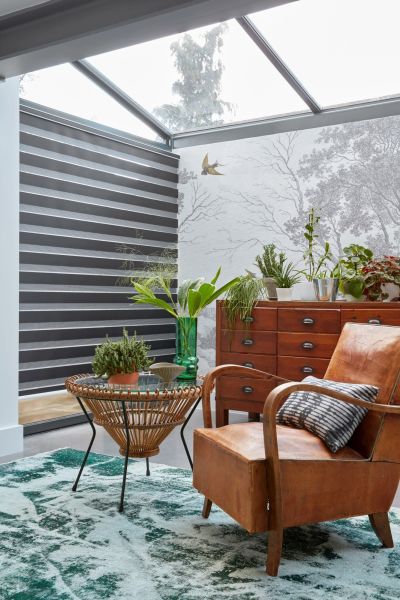 Striped dual-layer roller blind covering left glass wall, alternating dark and light horizontal bands, partially closed filtering daylight; sunroom with glass ceiling, leather armchair, rattan table, plants and wooden chest.