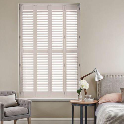 White four-panel plantation shutters covering tall window, horizontal louvers closed but slightly angled, filtering soft daylight; bedroom corner with upholstered bed, bedside table, lamp, vase of flowers and armchair.