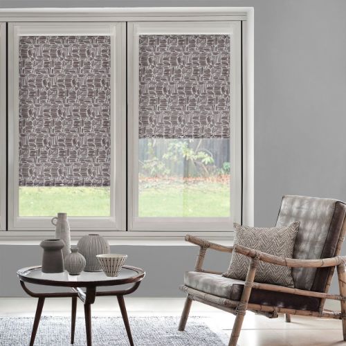 Patterned roller blinds (brown-and-white geometric) lowered halfway over two windows, partially filtering daylight; grey living room with a rattan leather armchair, coffee table, and grassy backyard view.