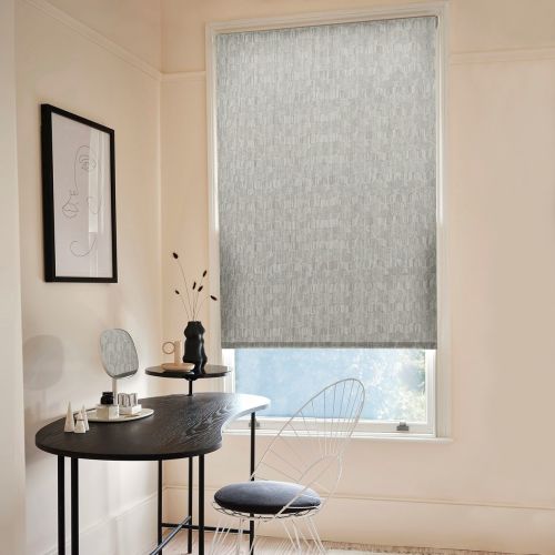 A grey patterned roller blind, lowered over a tall window, filtering daylight â€” in a pale minimalist dressing nook with a black desk, wire chair and framed line-art.