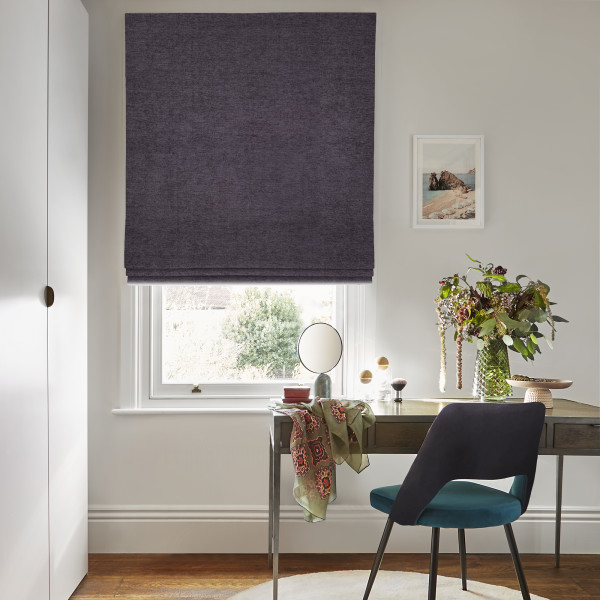 Roman fabric blind, deep plum, lowered with soft folds near the bottom; filtering daylight. Context: small home study — wooden desk with mirror, vase of foliage, teal-upholstered chair and wall art.