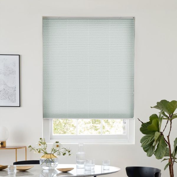 A pale green textured cellular blind, lowered over a window and filtering daylight, in a bright dining nook with round table, vase of flowers, framed art and a fiddle‑leaf fig.