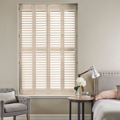 White wooden plantation shutters covering a tall inset window, closed with slatted louvers filtering soft daylight; bedroom setting with upholstered bed, bedside table, lamp and armchair in neutral tones.