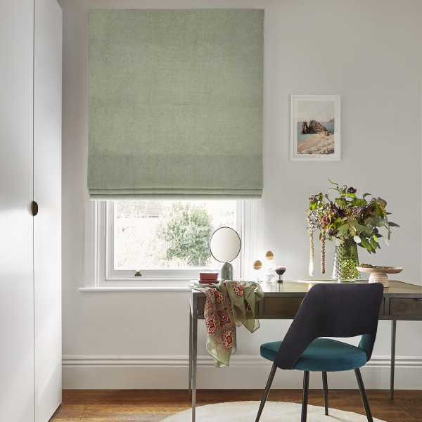 Sage-green fabric Roman blind, partially lowered with soft folds, filtering daylight through a white sash window; scene shows a wooden desk with teal chair, vase of flowers, mirror, pale walls.