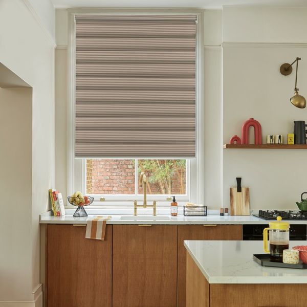 Beige horizontal pleated roller blind lowered over window, blocking most light with narrow stripes; modern kitchen sink area with brass faucet, wooden cabinets, marble countertops, island and shelving.