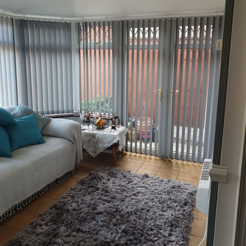 Vertical grey blinds cover bay windows and patio doors, mostly closed, filtering daylight through narrow slats. Cozy sunroom with sofa, teal cushions, lace-covered side table, tiled floor and shaggy rug.