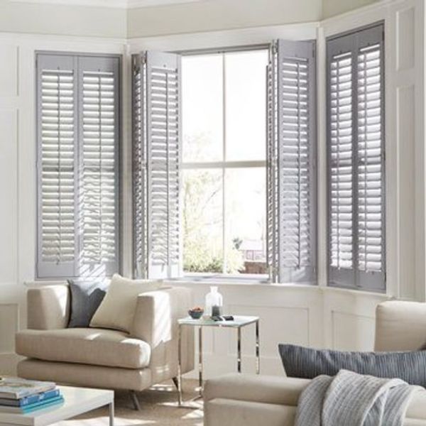 White wooden plantation shutters on a bay window; outer panels closed, center panels partially open, slats filtering bright daylight into a sunlit living room with beige sofas and side table.