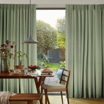 Floor-to-ceiling pleated sage-green curtains, partially parted, framing and softly filtering daylight onto a breakfast table set in a sunlit dining area opening onto a leafy garden.