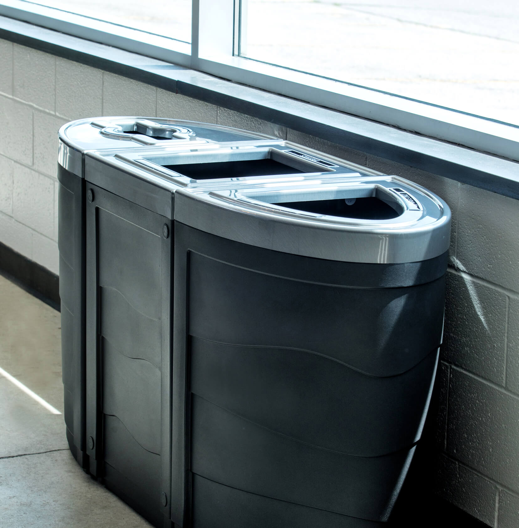 black plastic waste and recyclables containers with pewter patterned lids along a wall in front of windows at a municipal recreation facility