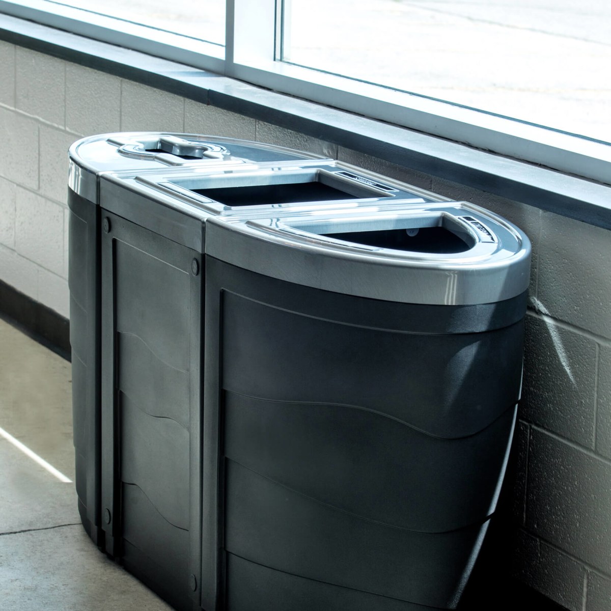 black plastic waste and recyclables containers with pewter patterned lids along a wall in front of windows at a municipal recreation facility
