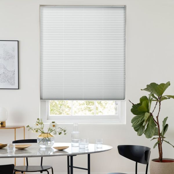 White pleated blind lowered over a recessed window, filtering soft daylight; dining table with glassware and vase, black chairs and a fiddle‑leaf fig plant in a bright minimalist room.