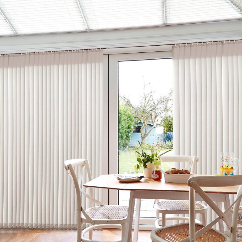 Cream vertical blinds covering patio doors, parted centrally to reveal a glass door; they hang in straight slats, softly filtering daylight in a bright dining nook overlooking a garden.