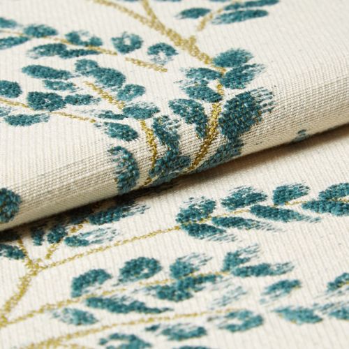Cream woven fabric with teal embroidered leaf motifs folded over itself, showing raised tufted stitching; macro close-up on a neutral surface with soft, diffuse lighting.