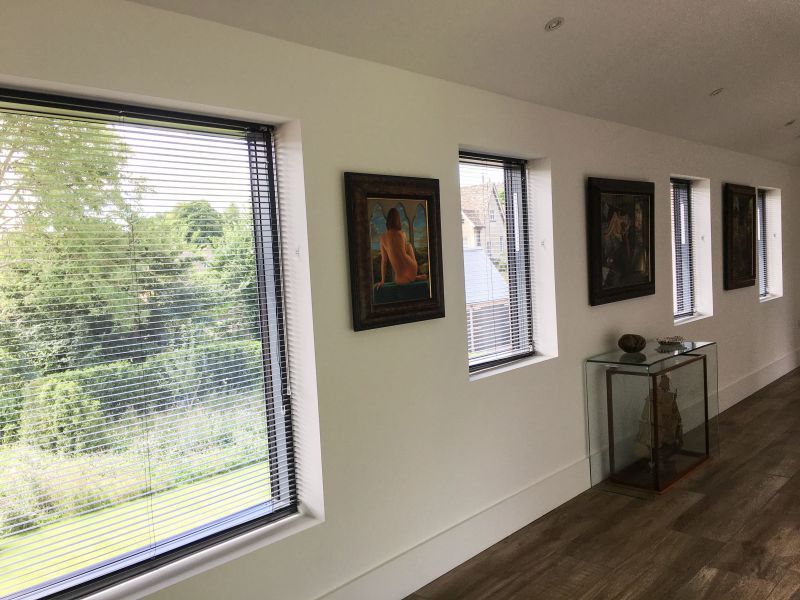 Horizontal dark Venetian blinds cover a series of deep-set windows, slats partially closed and filtering daylight; corridor-like gallery with white walls, framed paintings, a glass display case, and tree views.
