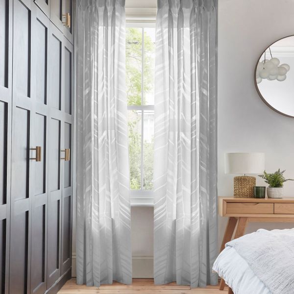 Sheer patterned floor-length curtains, slightly parted over a sash window, filtering soft daylight; bedroom with dark paneled wardrobe to the left, wooden bedside table, round mirror and greenery outside.