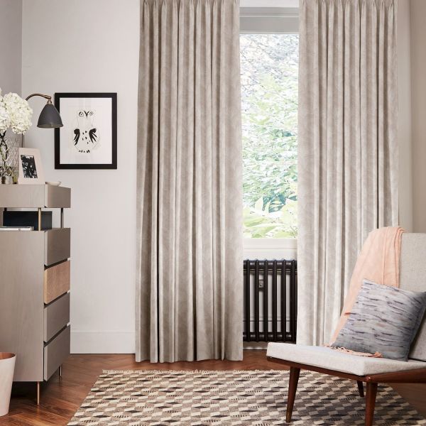 Beige patterned pleated curtains, hung floor-to-ceiling and drawn closed at sides leaving central pane exposed, filtering soft daylight; living-room setting with radiator beneath window, armchair, sideboard and patterned rug.