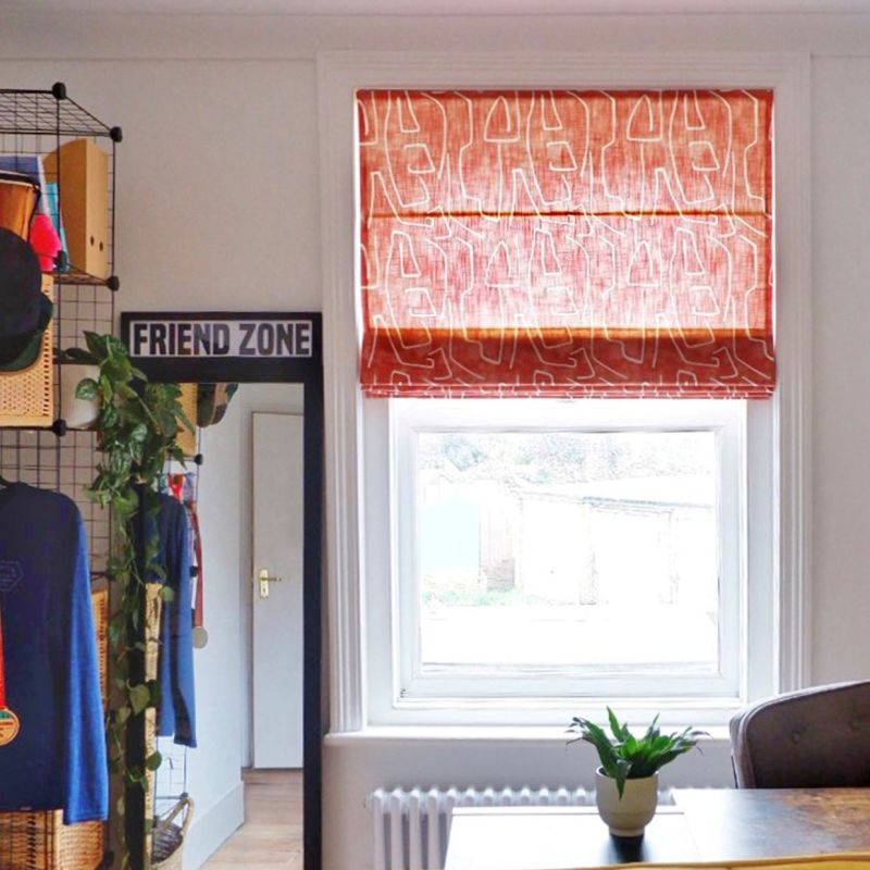 Red-orange Roman blind with white abstract-line pattern, partially lowered and filtering daylight over a sash window; bright room with radiator, desk and potted plant, wire shelving and mirror labeled FRIEND ZONE.